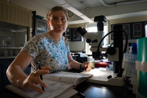 Photo of Catherine (Catrina) Nowakowski in front of microscope in lab